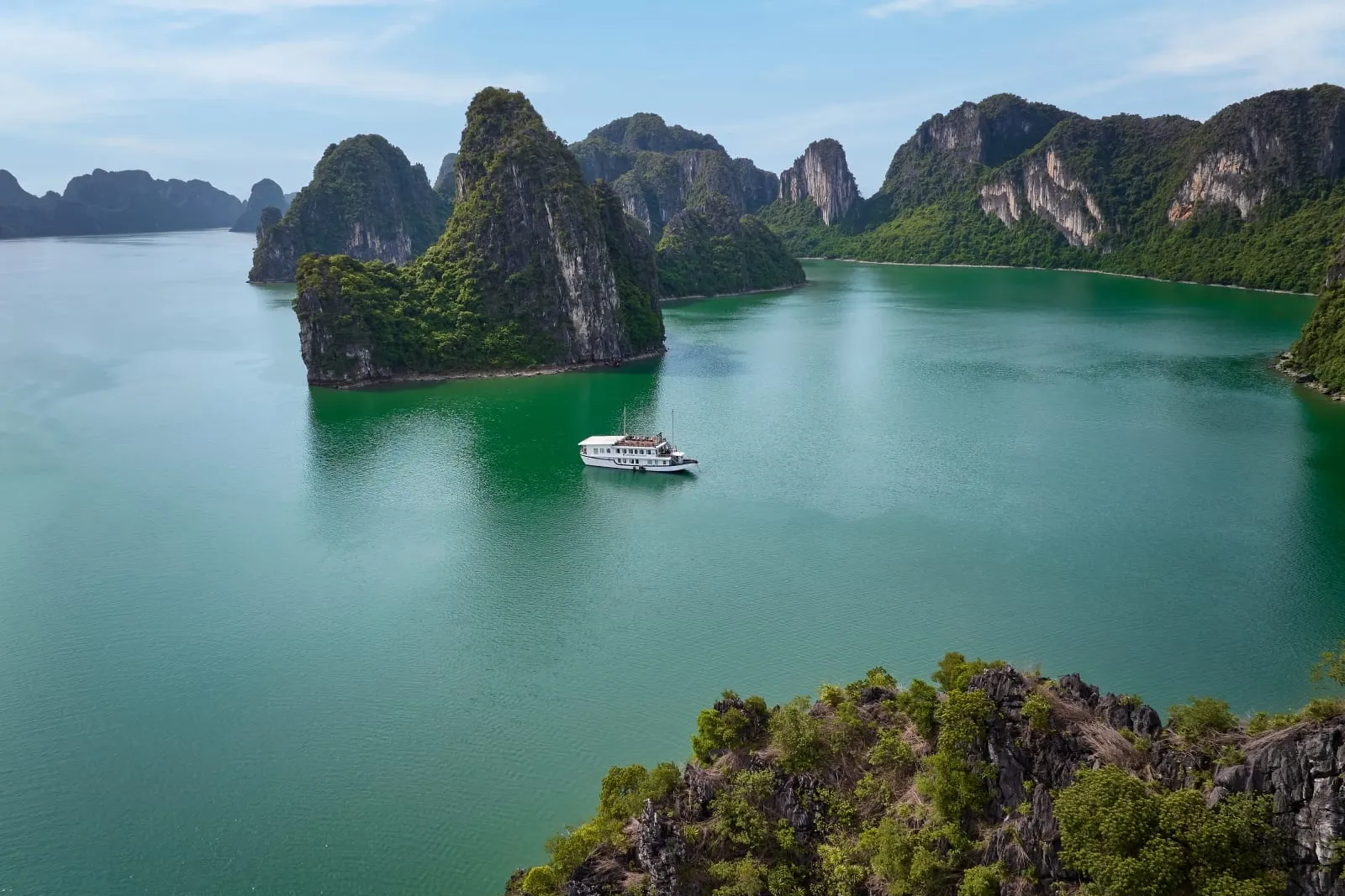 An elevated view of emerald-green water surrounded by tree-covered limestone karst islands, with a single small white boat anchored in the middle.