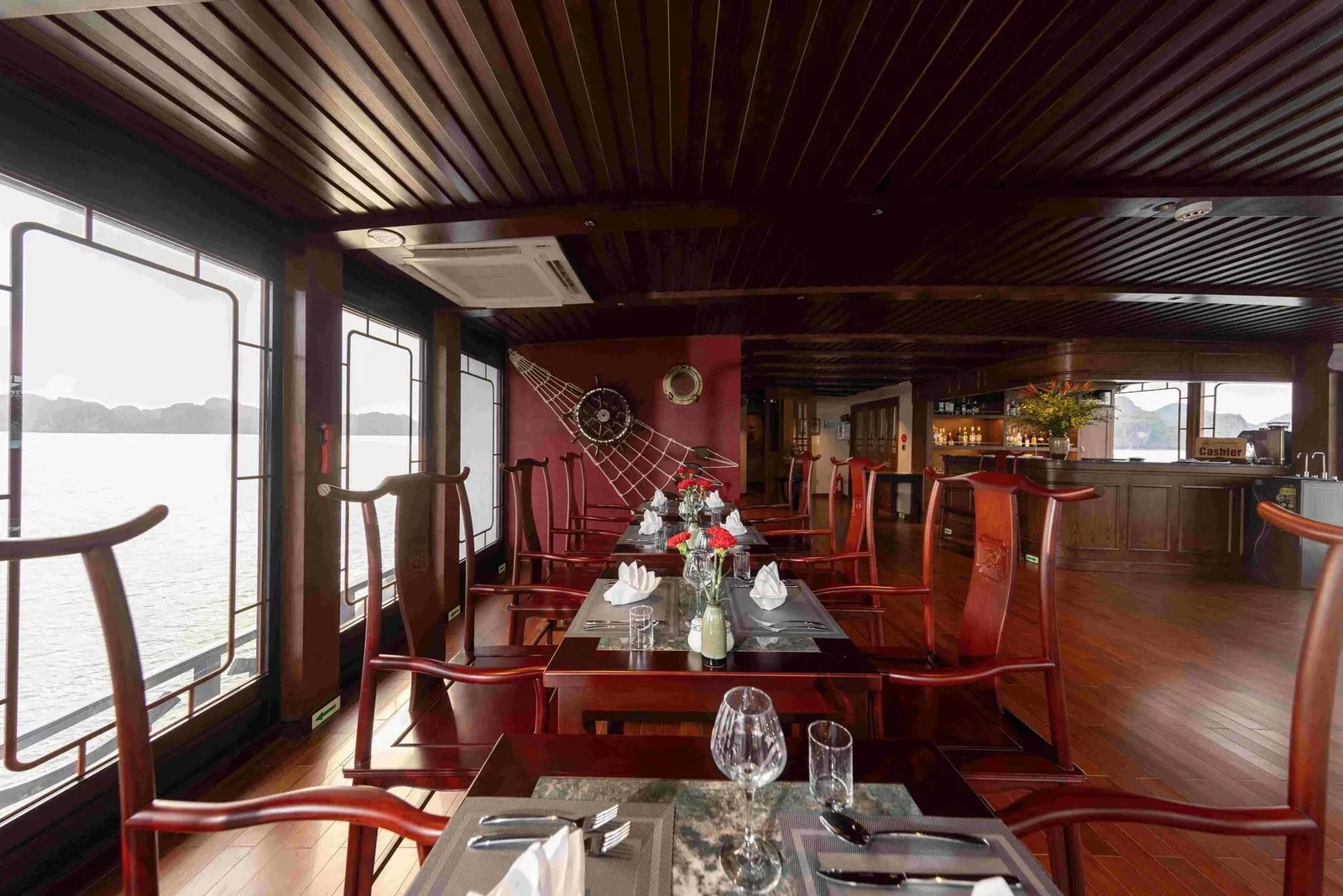 A wood-paneled dining room on a boat with set tables, red carpentry chairs, a ship's wheel and net decoration on the wall, a bar with a 'Cashier' sign, and large windows showing calm water and distant limestone hills.