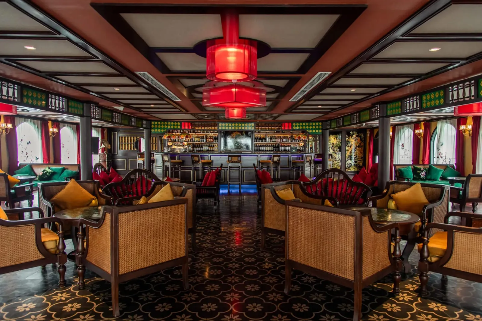 An interior lounge and bar with rattan and dark wood armchairs, gold and red cushions, red pendant lamps, a patterned tile floor, and a bar with bottles at the far end.
