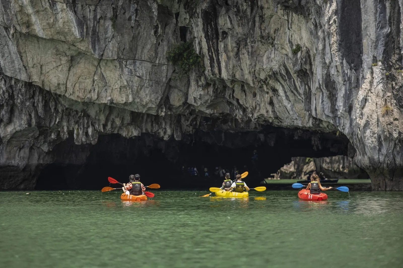 Three kayaks with paddlers in life vests approach a large dark limestone cave opening over green water.