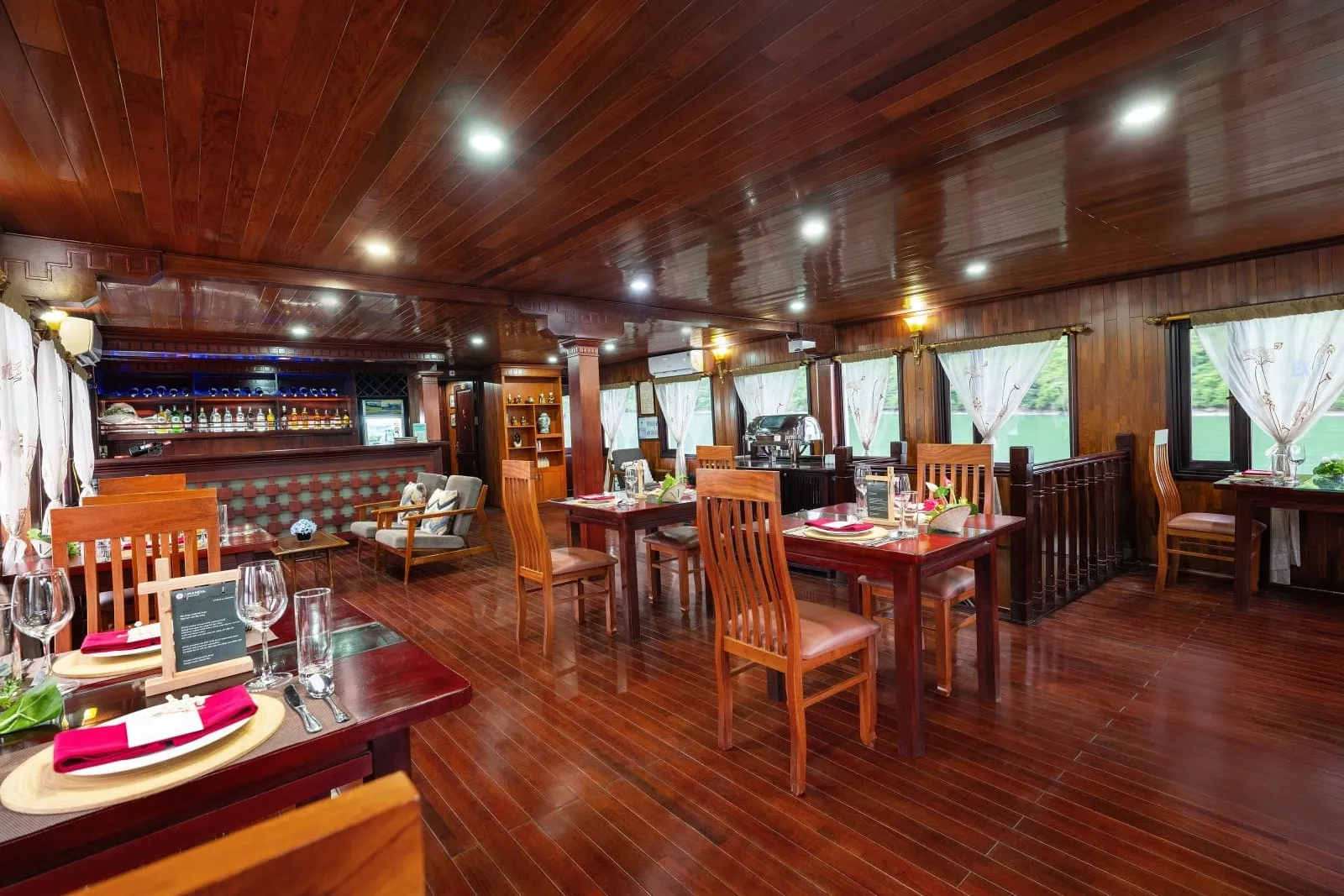 A wood-paneled boat dining room with set tables, red napkins, a bar with bottles at the back, and windows showing green water and tree-covered karst hillsides.