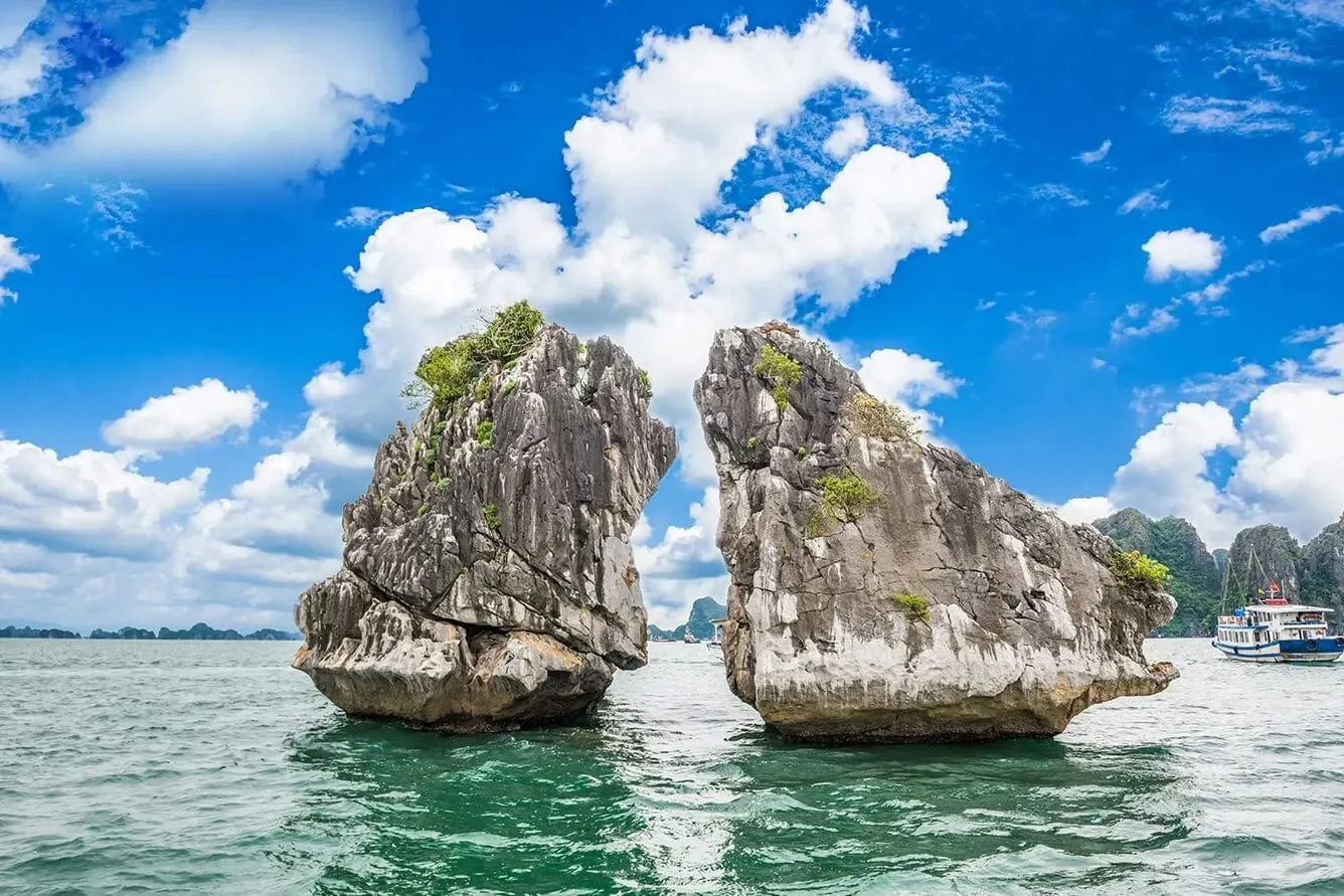 Two large limestone karst formations rise from green water under a blue sky with white clouds, with cruise boats visible in the background.