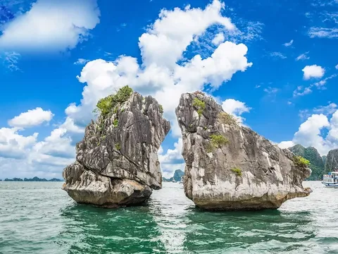 Two large limestone karst formations rise from green water under a blue sky with white clouds, with cruise boats visible in the background.