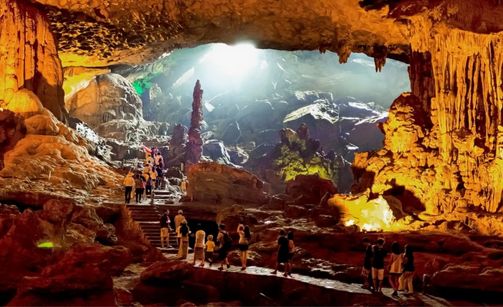 Tourists walk through a large illuminated cave with stalactites and stalagmites, with wooden stairs and platforms throughout.