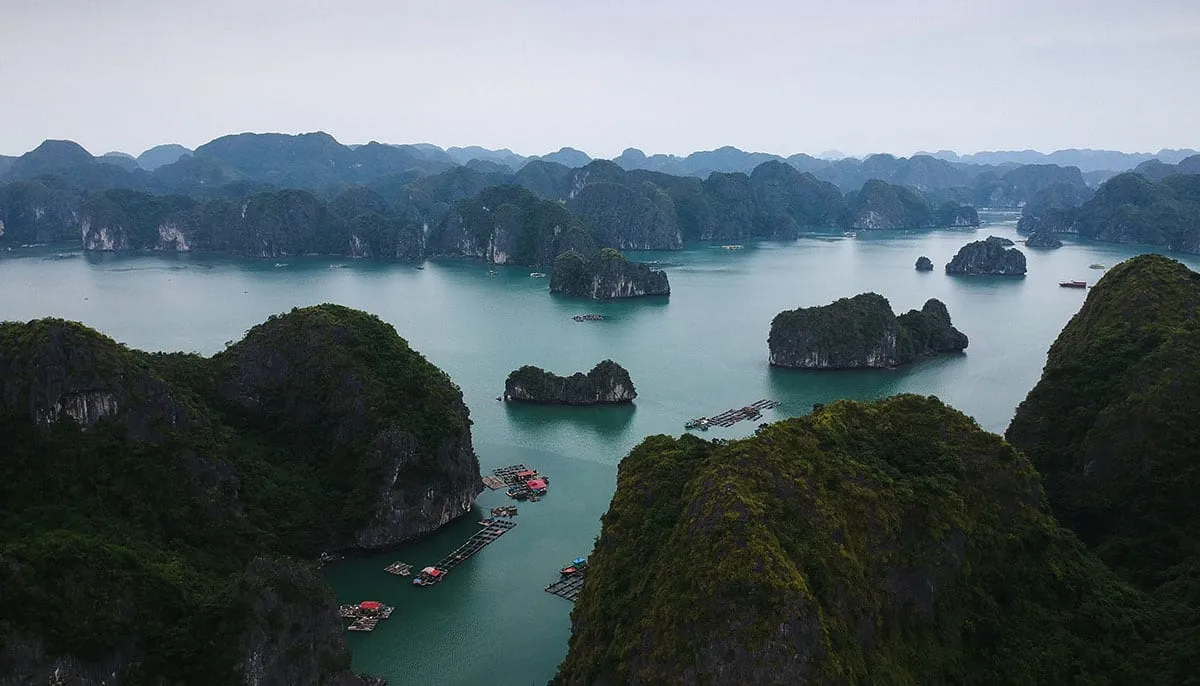 An aerial view of limestone karst islands rising from turquoise water with a floating village visible in the middle distance.
