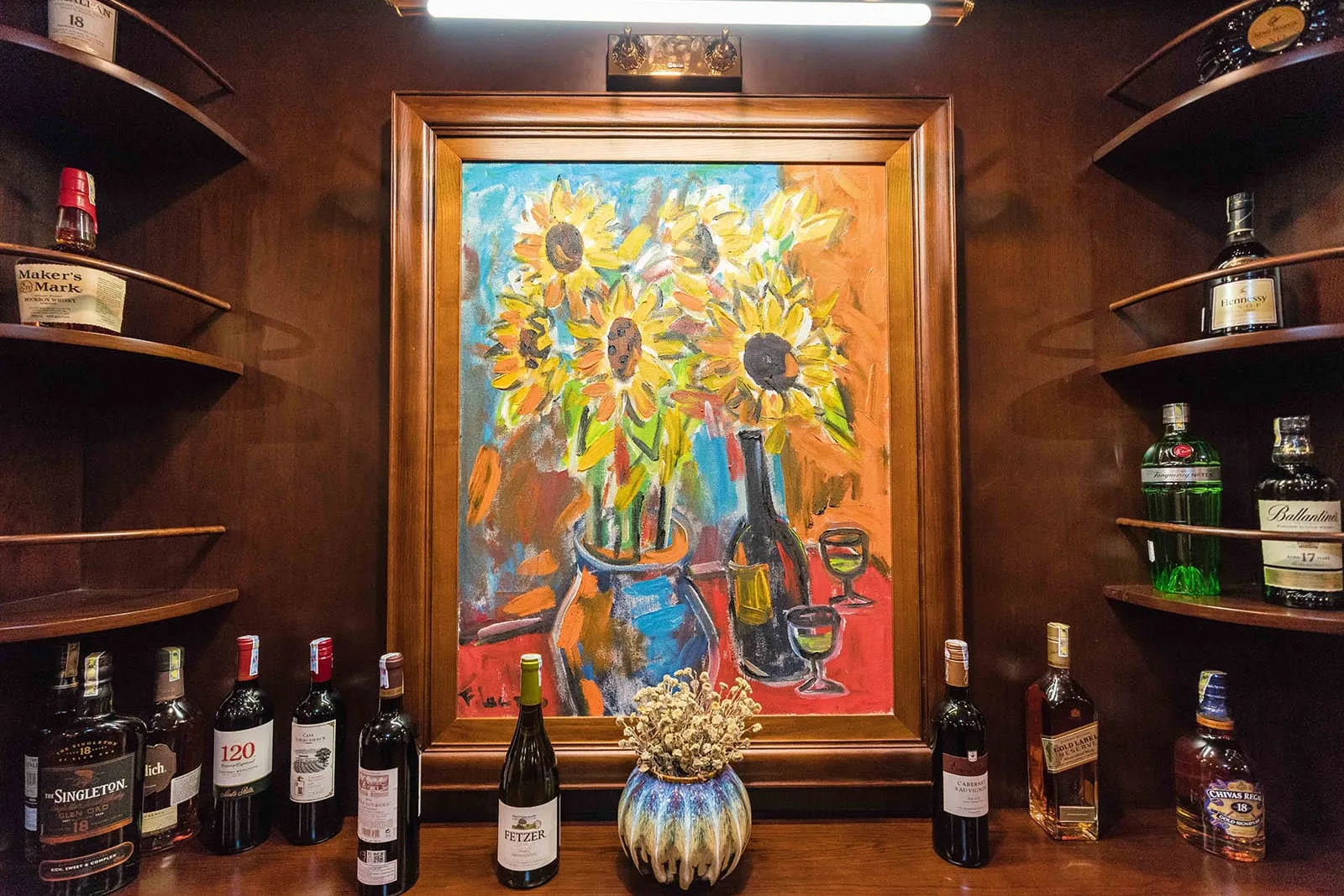 A wooden bar display with liquor bottles on shelves flanking a framed sunflower painting, with wine bottles and a decorative vase arranged below.