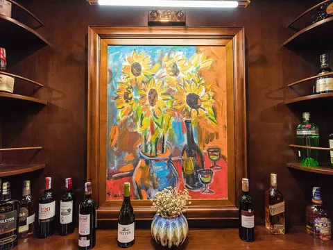 A wooden bar display with liquor bottles on shelves flanking a framed sunflower painting, with wine bottles and a decorative vase arranged below.