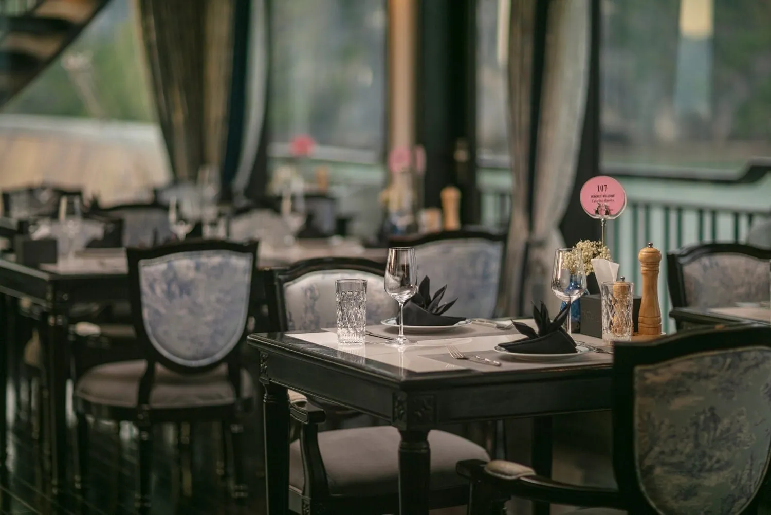 Interior of a dining room with set tables, dark wooden chairs with blue floral upholstery, folded black napkins, glasses, a pepper mill and a pink table number card, with blurred windows and a green railing in the background.