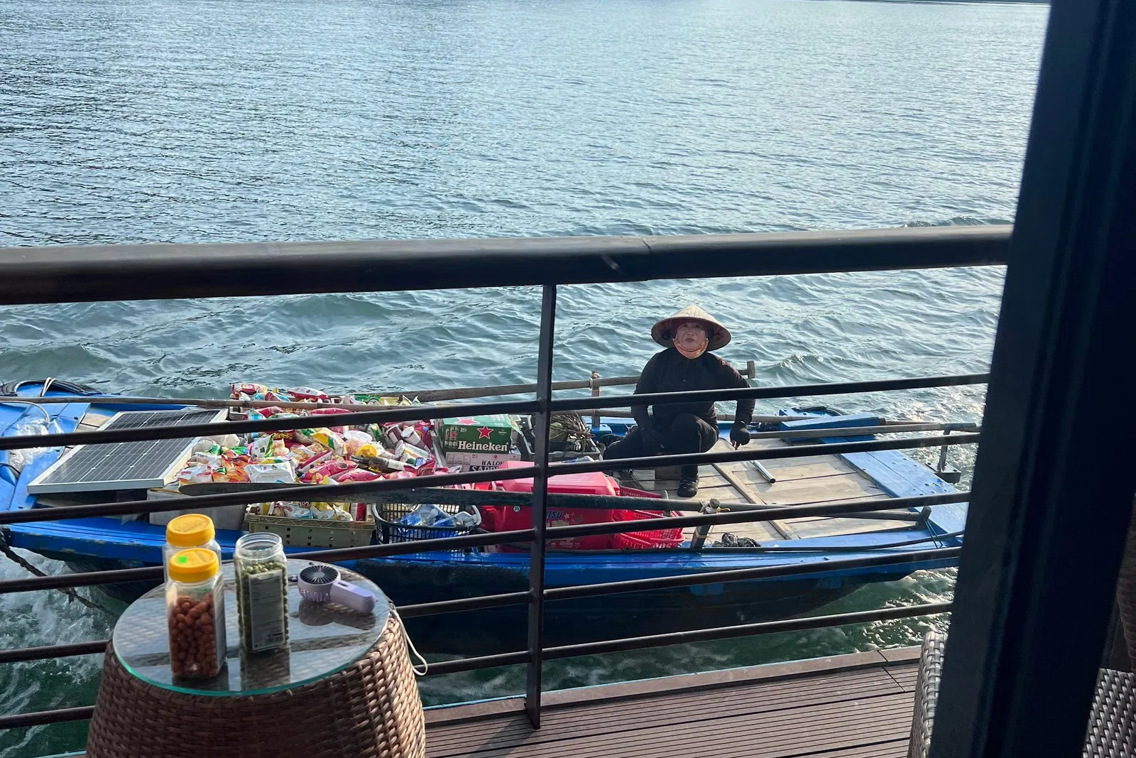 A floating vendor pulls alongside in a blue boat stacked with snacks, seen from the deck table
