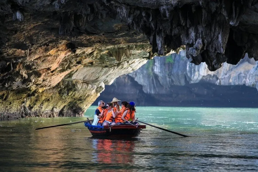 A group of tourists in orange life jackets sit in a small rowboat inside a limestone cave with turquoise water and dramatic rock formations.