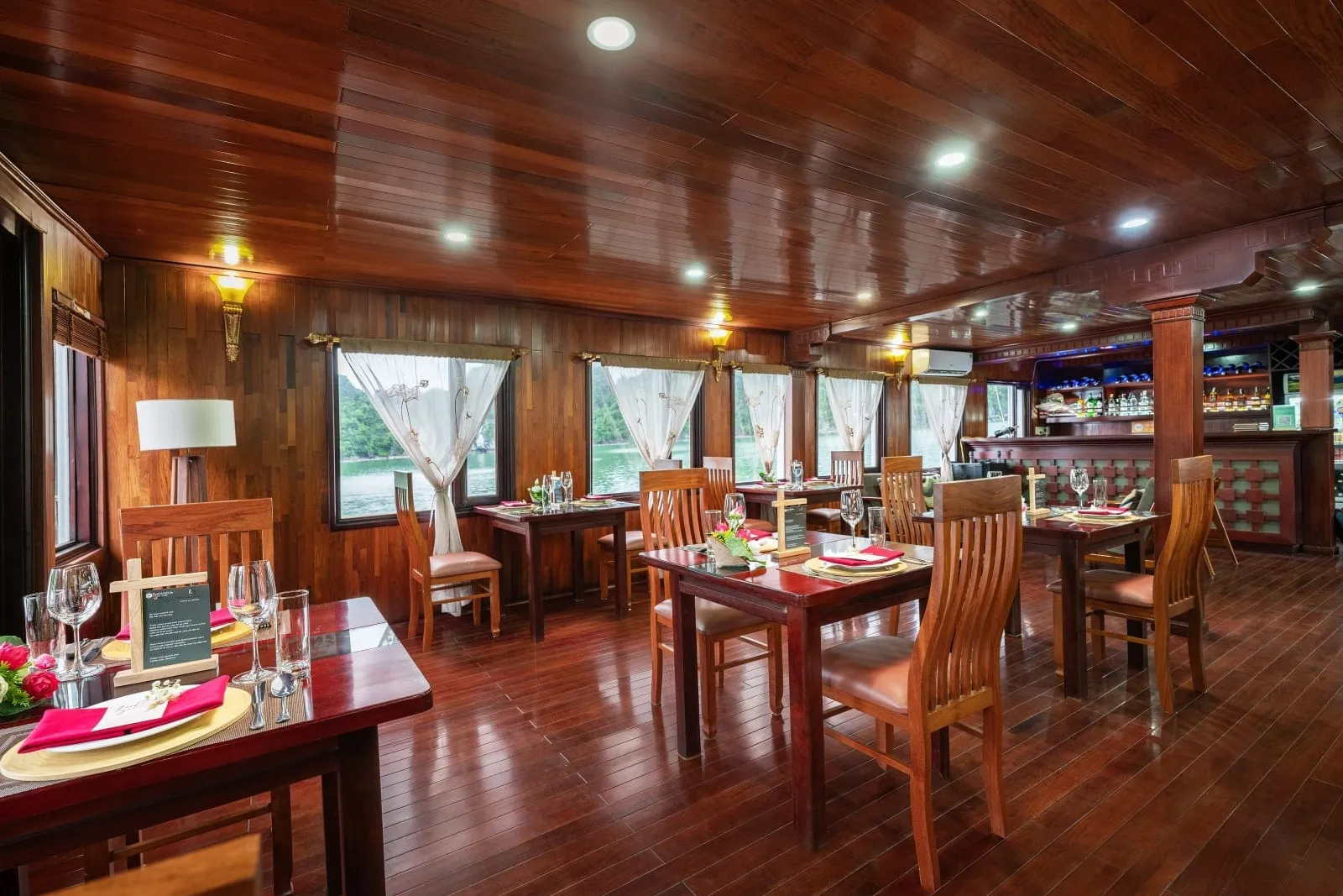 A wood-paneled dining room on a boat with set tables, red napkins, wooden chairs and a bar at the back, with windows showing limestone karst islands and green water.