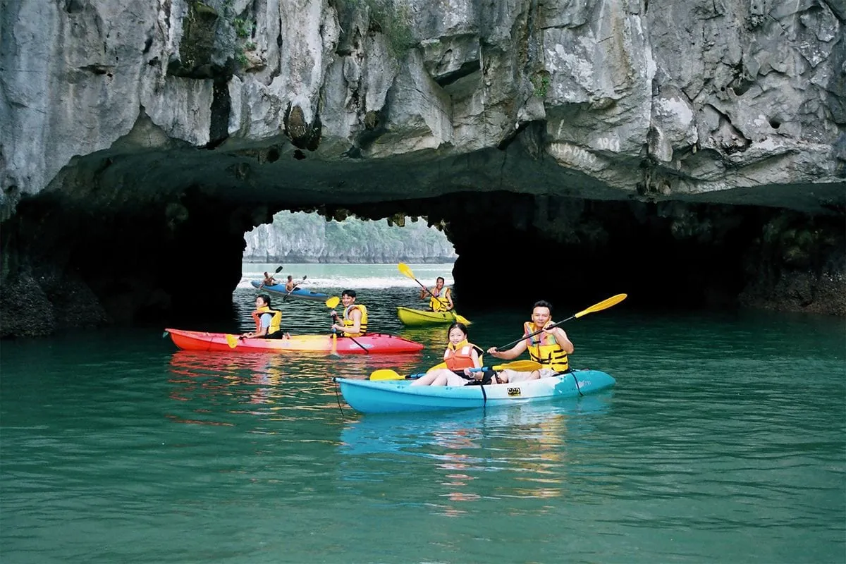 Multiple kayakers in colorful kayaks paddling through a cave opening with limestone karst formations and emerald water.