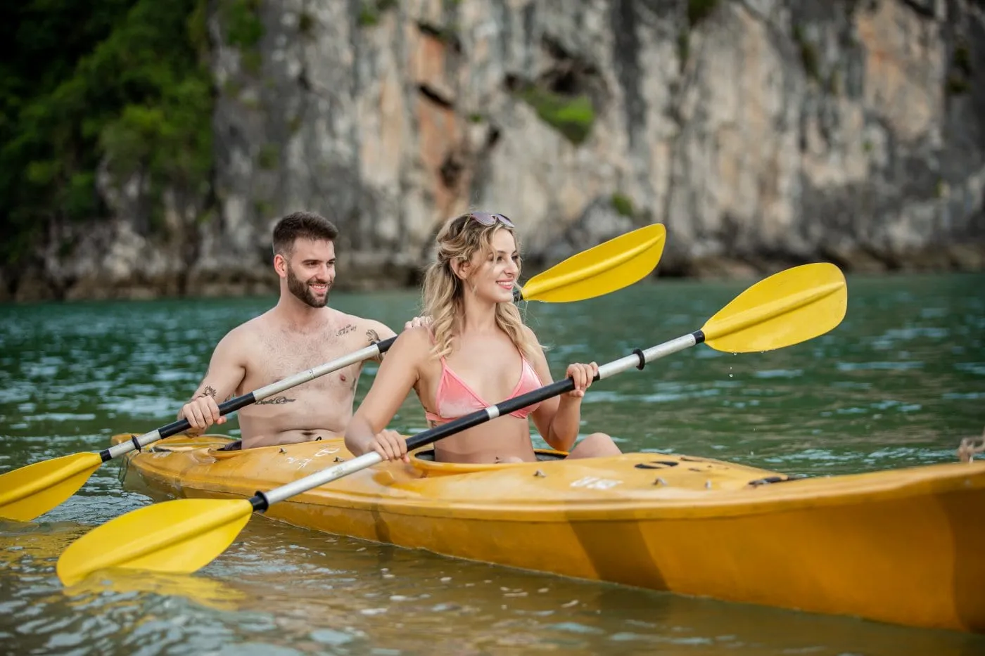 Two people kayaking in a yellow tandem kayak on calm water with tall limestone karst cliffs in the background.