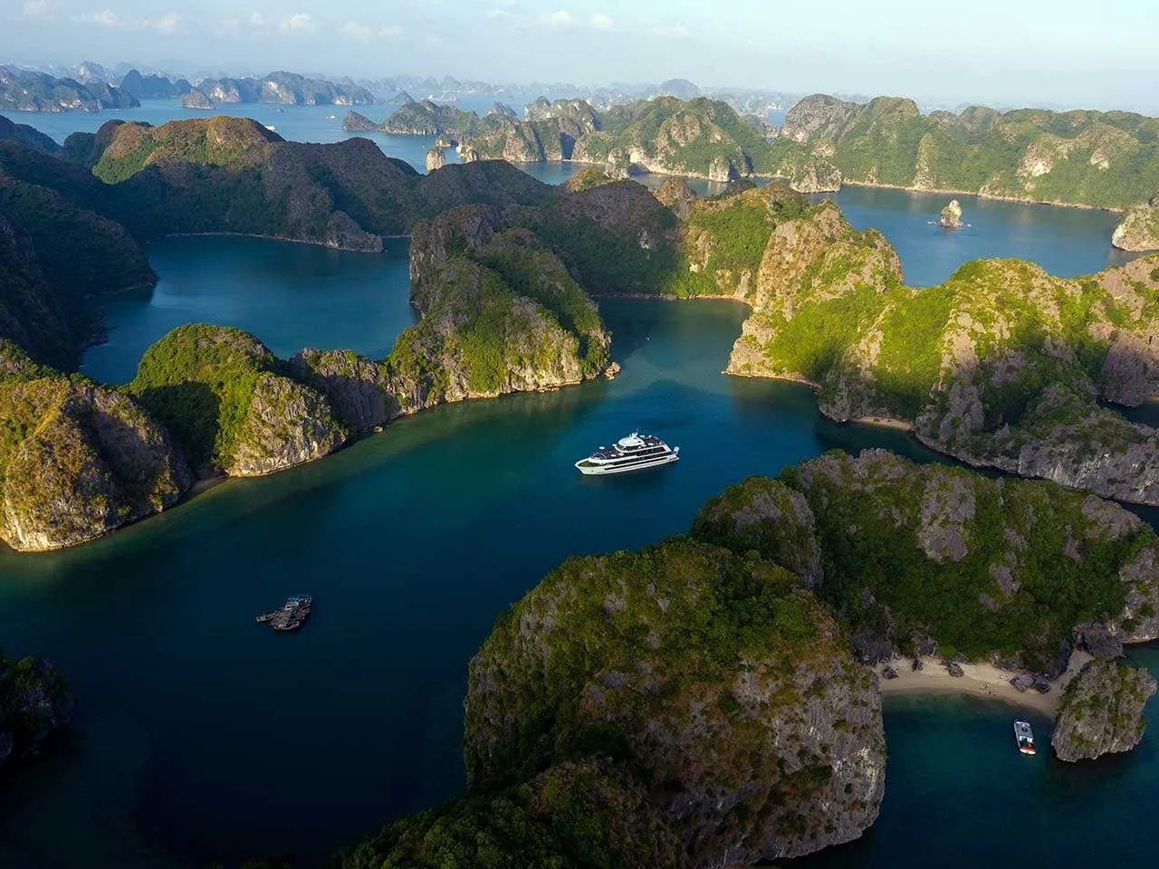 Aerial view of limestone karst islands covered in greenery with a small white multi-deck cruise ship in the middle of turquoise water, plus a small fishing boat and a speedboat near a beach.