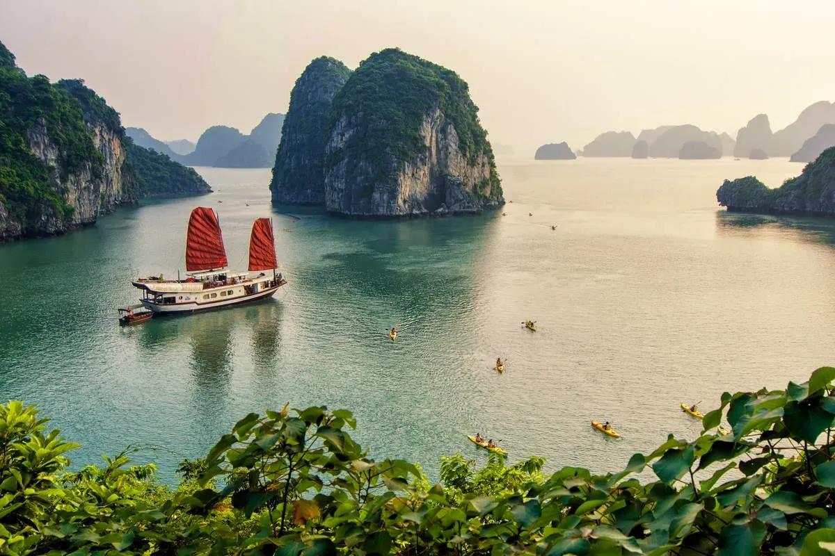 A traditional junk boat with red sails floats in calm green water surrounded by tall limestone karsts, with kayakers visible in the foreground and foliage framing the bottom of the shot.