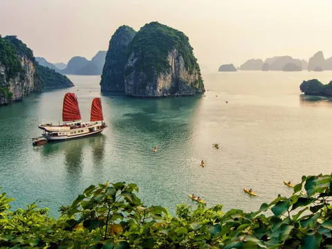 A traditional junk boat with red sails floats in calm green water surrounded by tall limestone karsts, with kayakers visible in the foreground and foliage framing the bottom of the shot.