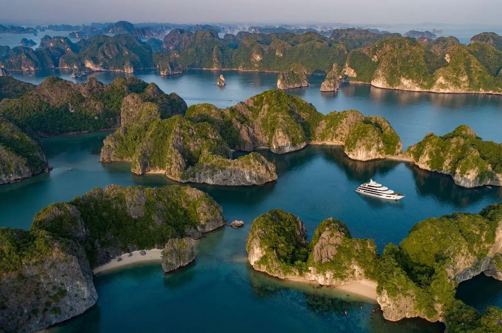 Aerial view of limestone karst islands covered in green vegetation with a small white multi-deck cruise ship anchored in teal water between them.