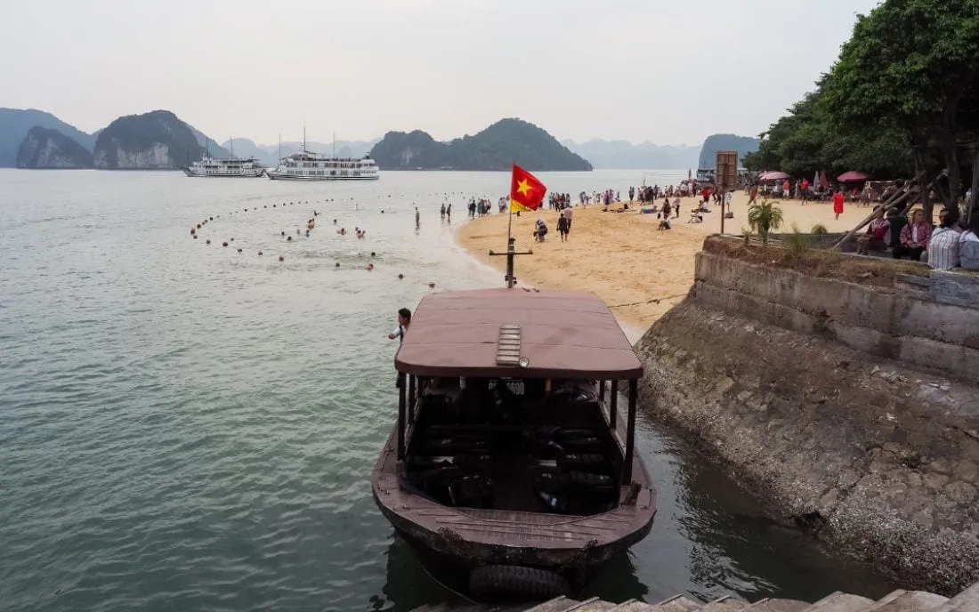 A beach with limestone karst islands in the background, cruise ships anchored offshore, people on the sand, a Vietnamese flag, and a small tender boat docked in the foreground.