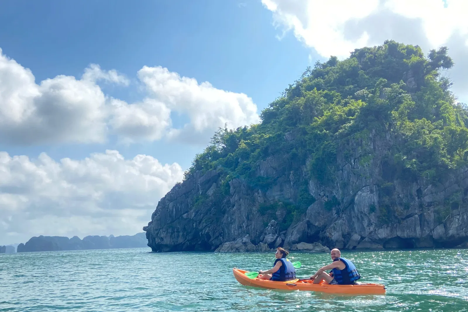 Two people in blue life vests paddle an orange tandem kayak on green water beside a steep, tree-covered limestone island, with more karst islands in the distance under a blue sky with clouds.