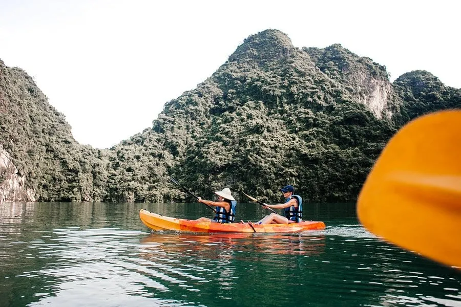 Two people in life jackets paddle an orange kayak on calm water with limestone karst mountains in the background.