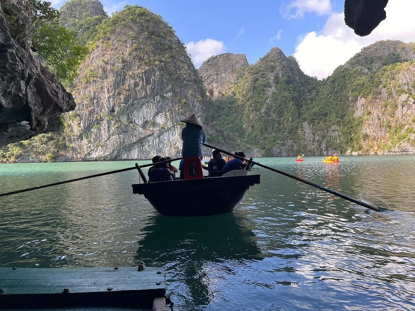 A rower in a conical hat stands in a small wooden rowing boat carrying passengers in life jackets across green water between limestone karst cliffs, with kayakers in the distance and a wooden platform in the foreground.