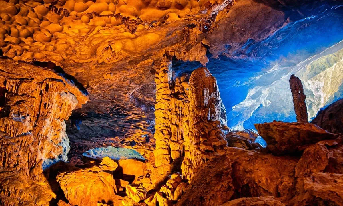 Interior of an illuminated cave with orange-lit stalactites and stalagmites and blue-lit ceiling openings.