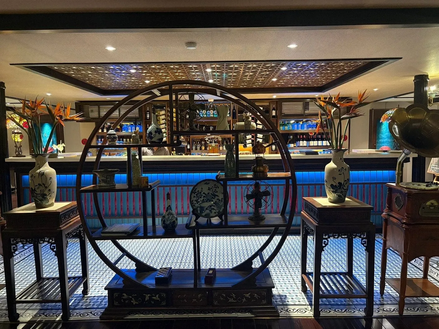 Interior bar lounge with a round wooden display shelf holding blue-and-white porcelain, flanking tables with vases of bird-of-paradise flowers, a gramophone at right, a lit bar with bottles behind, and a patterned blue-and-white tile floor.