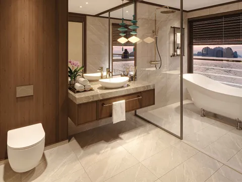 A luxury bathroom with marble finishes, a freestanding tub, and large windows showing karst limestone formations typical of Ha Long Bay at sunset.