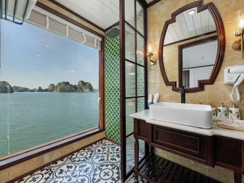 A boat cabin bathroom with a large window showing limestone karsts rising from green water, featuring a vessel sink, decorative mirror, patterned tile floor, and yellow marble walls.