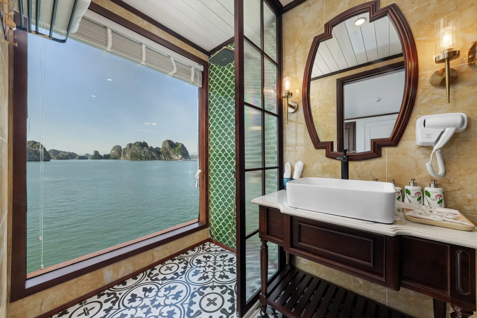 A boat cabin bathroom with a large window showing limestone karsts rising from green water, featuring a vessel sink, decorative mirror, patterned tile floor, and yellow marble walls.