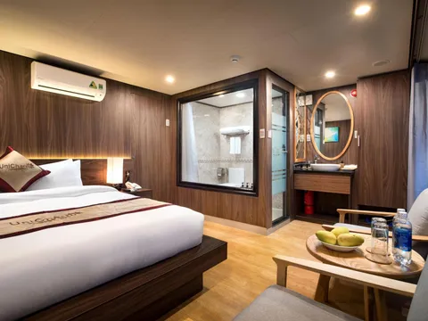 A wood-panelled cruise cabin with a double bed, a glass-walled bathroom, a small sink area with an oval mirror, and two armchairs around a round table with mangoes and water bottles, with 'UniCharm' branding on the pillow and bed runner.