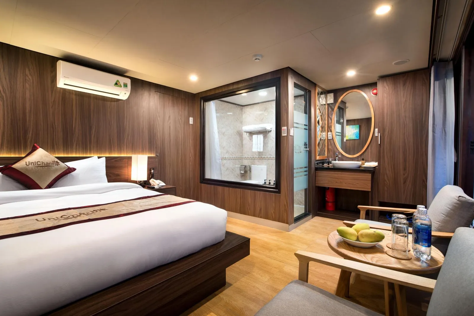 A wood-panelled cruise cabin with a double bed, a glass-walled bathroom, a small sink area with an oval mirror, and two armchairs around a round table with mangoes and water bottles, with 'UniCharm' branding on the pillow and bed runner.