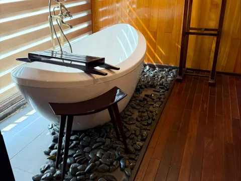 A white freestanding oval bathtub with a wooden caddy sits on a bed of black pebbles in a wood-paneled room with dark plank flooring, a wooden valet stand with a towel, and sunlight coming through horizontal window blinds.