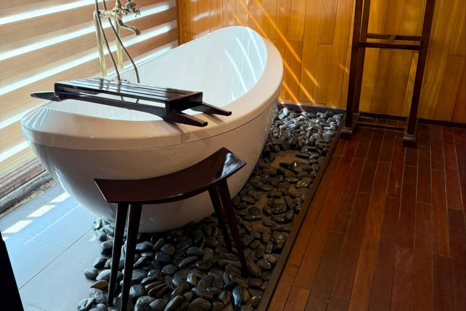 A white freestanding oval bathtub with a wooden caddy sits on a bed of black pebbles in a wood-paneled room with dark plank flooring, a wooden valet stand with a towel, and sunlight coming through horizontal window blinds.