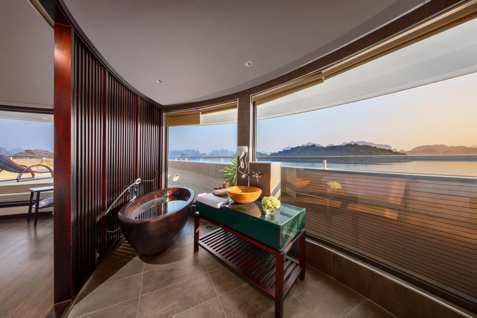 A curved cabin bathroom with a dark wooden freestanding tub and an orange vessel sink on a green stone vanity, with floor-to-ceiling windows overlooking a bay with limestone karst islands at sunset.