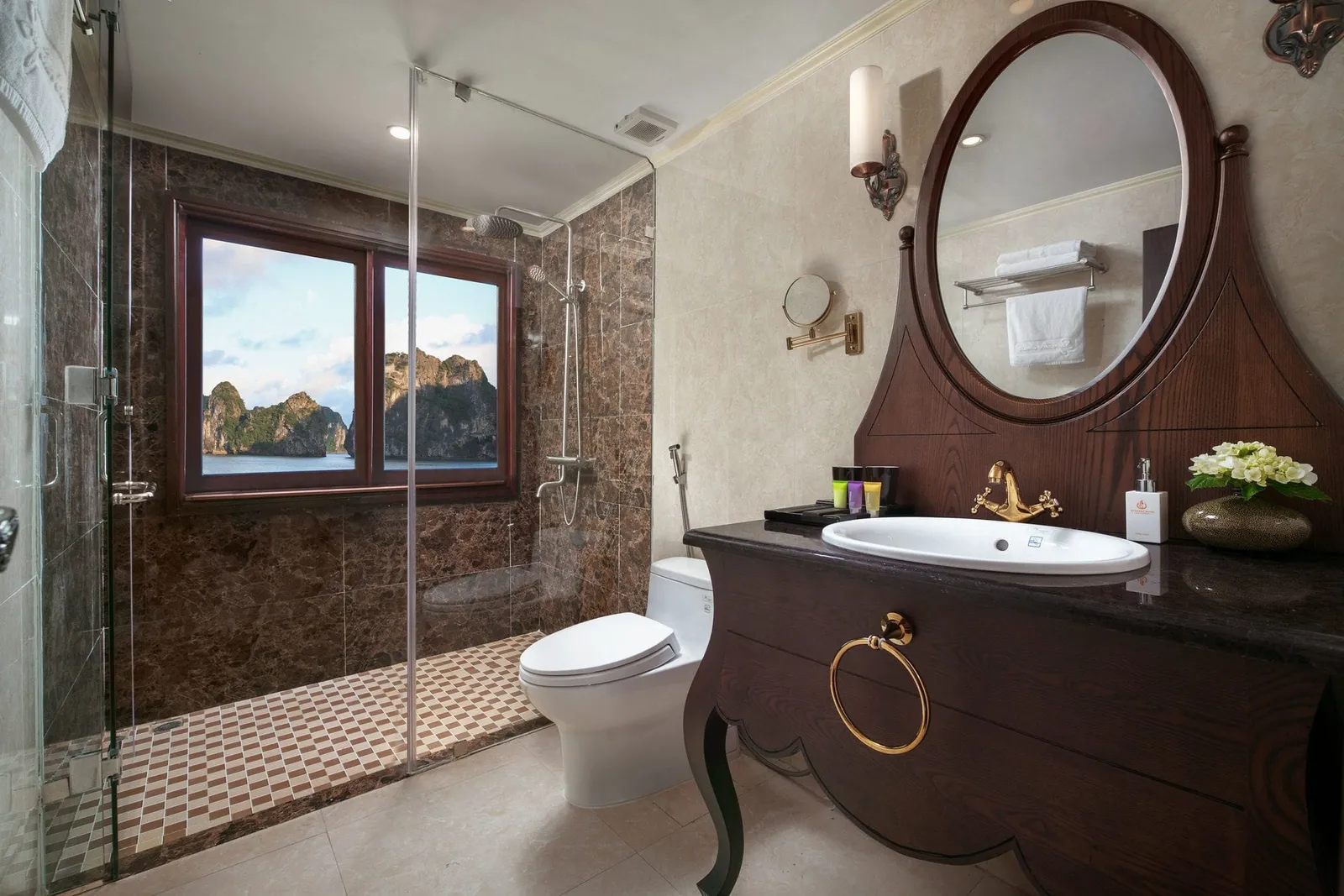 A cabin bathroom with a glass-enclosed marble shower, a toilet, and a dark wooden vanity with an oval mirror and a gold faucet, with a window showing limestone karst islands over water.