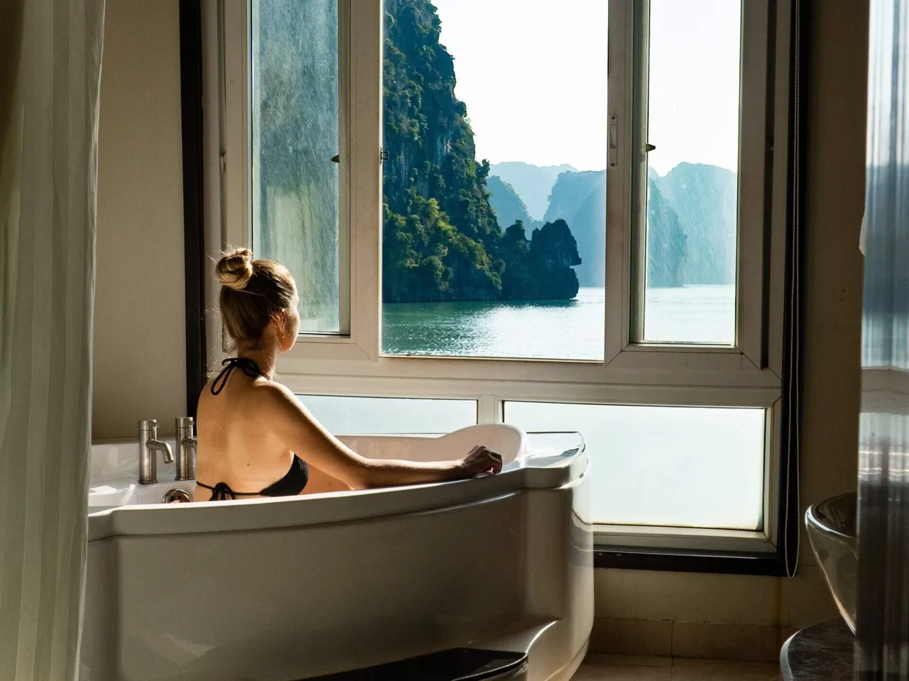 A person in a bathtub inside a cabin with windows showing limestone karst islands and calm water outside.