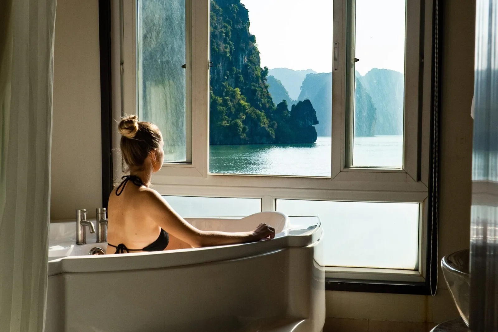 Guest in a freestanding bath looking out through the cabin window at karst cliffs across the water