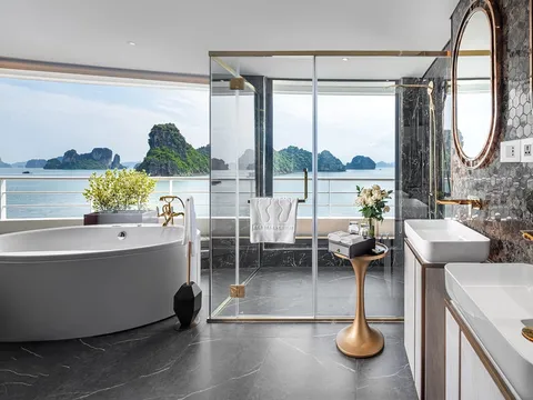 A luxury bathroom with a freestanding tub, dual sinks with gold fixtures, and floor-to-ceiling glass doors opening to a balcony overlooking limestone karsts and calm waters.