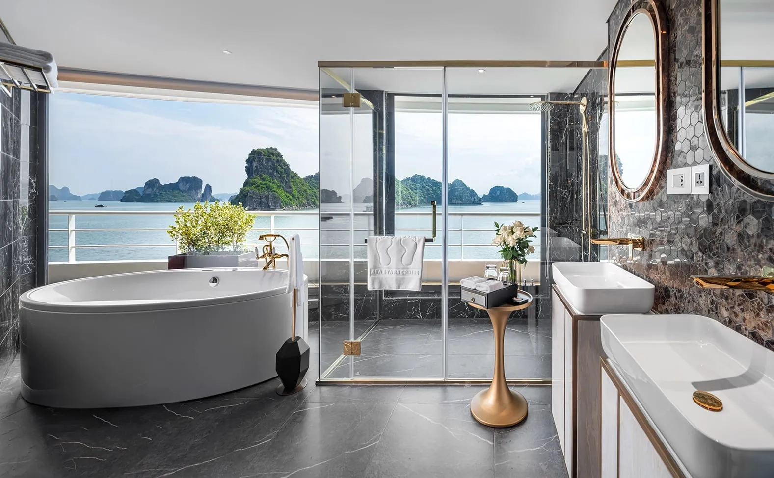 A luxury bathroom with a freestanding tub, dual sinks with gold fixtures, and floor-to-ceiling glass doors opening to a balcony overlooking limestone karsts and calm waters.