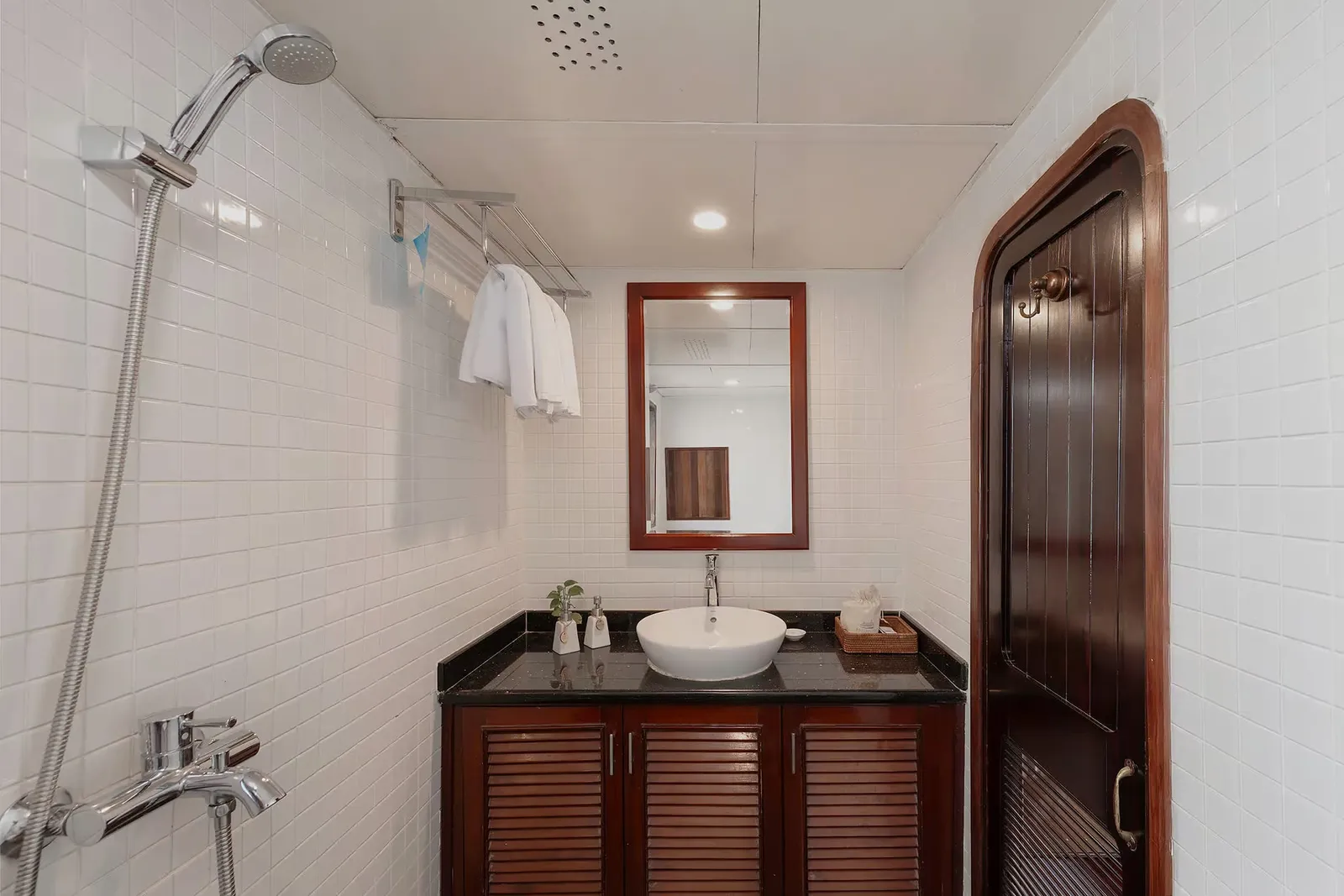 A small bathroom with white tiled walls, a handheld shower, a dark wood vanity with a black countertop and white vessel sink, a framed mirror, towels on a rack, and an arched dark wood door.