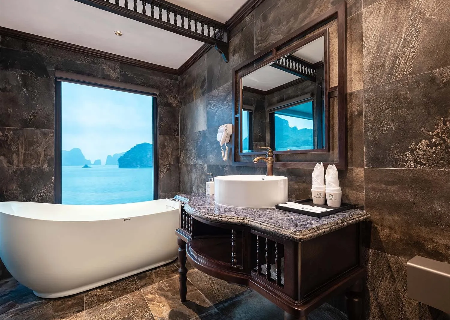 A dark-tiled luxury bathroom on a cruise boat with a freestanding white bathtub beside a large window overlooking karst limestone islands, a vessel sink on a dark wood vanity with branded toiletry bottles, and a wall-mounted hair dryer.