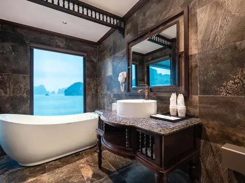 A dark-tiled luxury bathroom on a cruise boat with a freestanding white bathtub beside a large window overlooking karst limestone islands, a vessel sink on a dark wood vanity with branded toiletry bottles, and a wall-mounted hair dryer.