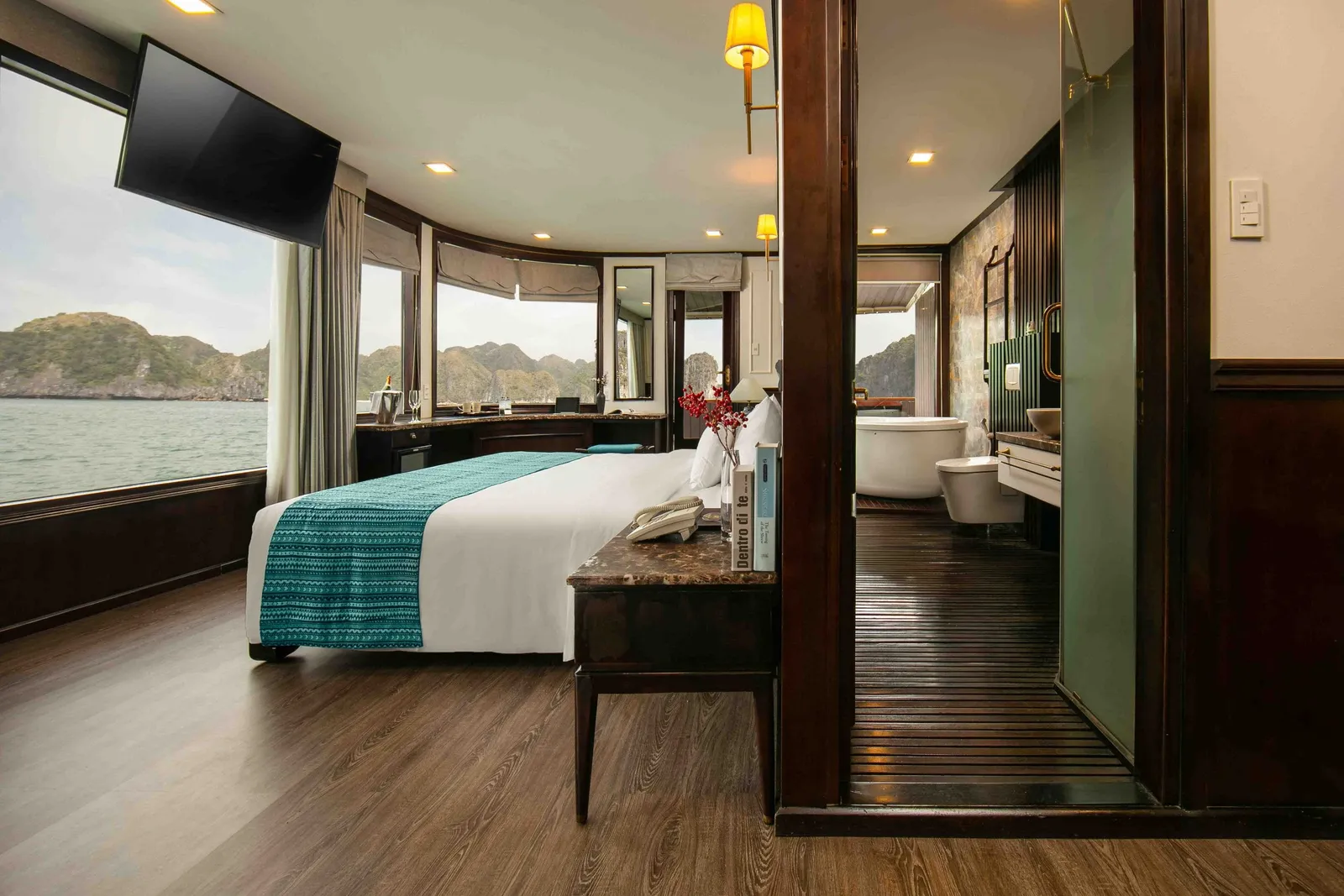 A cruise ship cabin interior with a bed, curved windows showing karst limestone islands and water, and an ensuite bathroom visible through an open door.