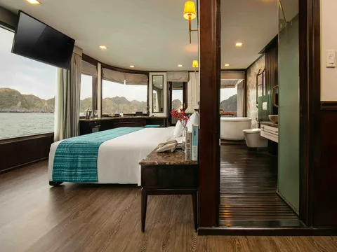 A cruise ship cabin interior with a bed, curved windows showing karst limestone islands and water, and an ensuite bathroom visible through an open door.