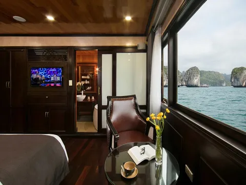 A luxury boat cabin interior with large windows showing limestone karsts and turquoise water outside, featuring a leather chair, coffee table with flowers and an open book, bed, TV, and visible bathroom.