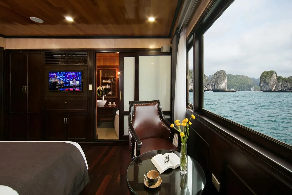 A luxury boat cabin interior with large windows showing limestone karsts and turquoise water outside, featuring a leather chair, coffee table with flowers and an open book, bed, TV, and visible bathroom.