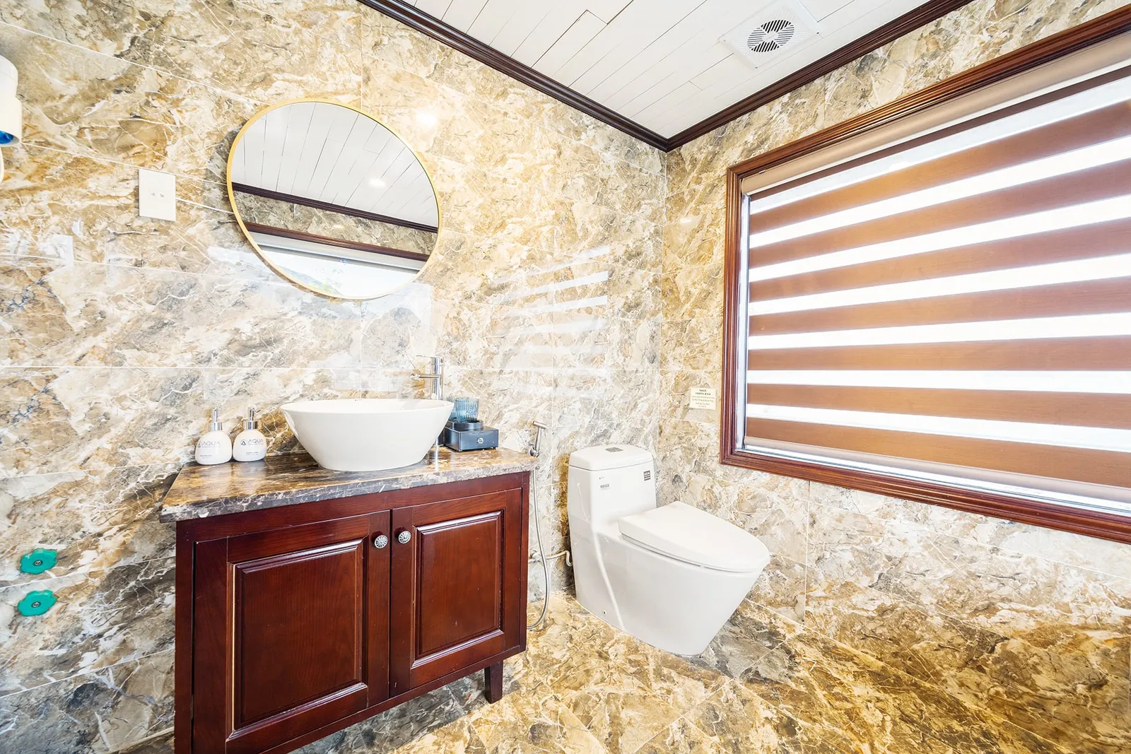 A bathroom with marble-look tiled walls and floor, a dark wood vanity with a white vessel sink, a round gold-framed mirror, a white toilet, and a window with a striped brown and white zebra blind; the soap dispensers carry small printed labels.