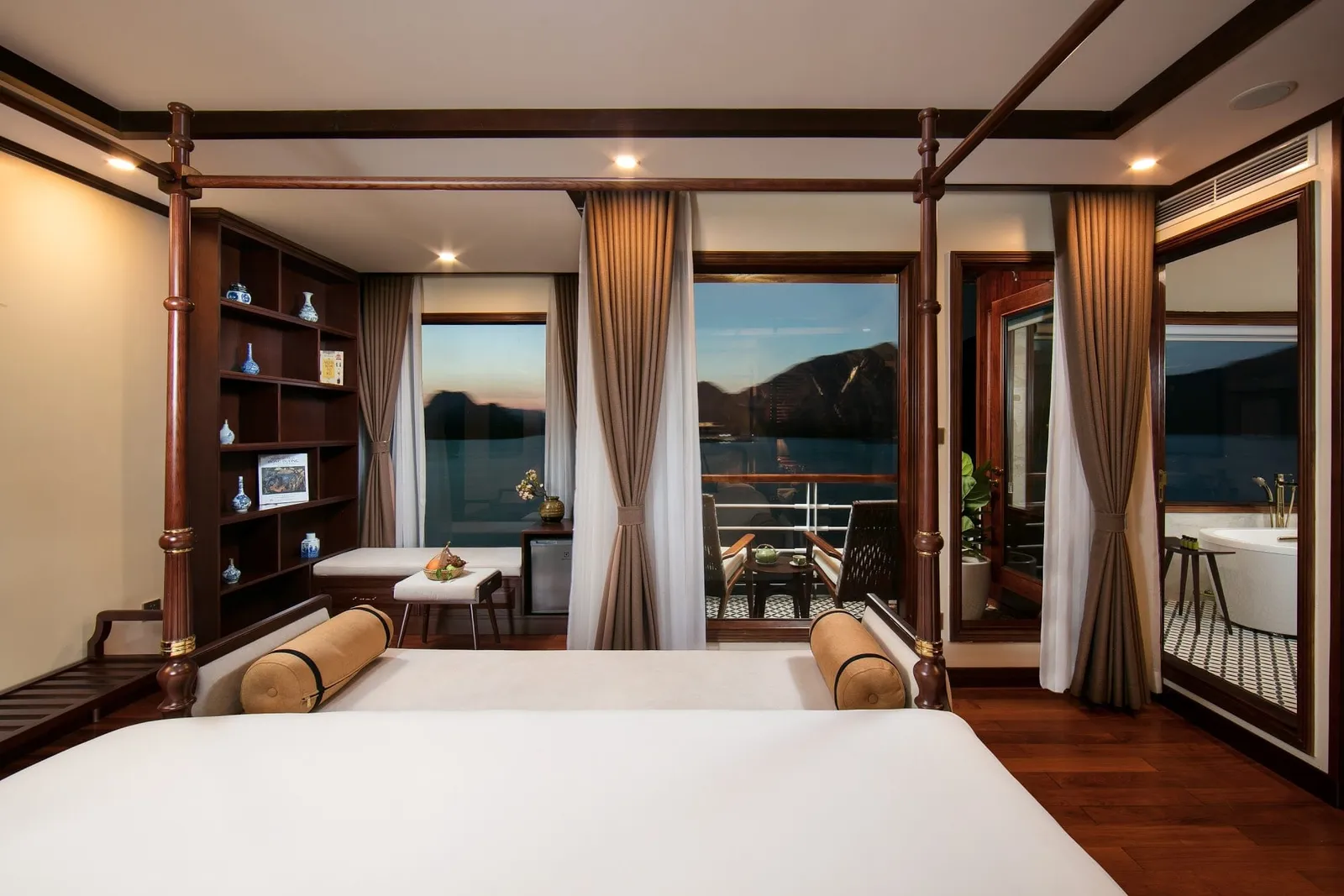 A cabin with a dark wood four-poster bed, shelves with blue-and-white vases, and a private balcony with chairs overlooking a bay with karst-like hills at dusk, plus a bathtub visible through a side door.