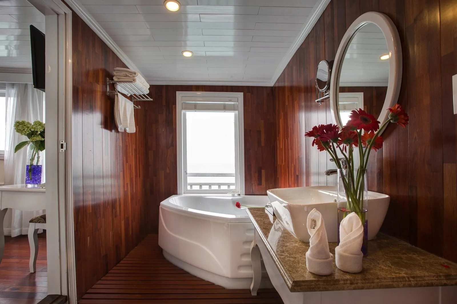 A wood-paneled cabin bathroom with a corner bathtub, vessel sink on a marble counter with red gerbera flowers and folded towels, a round mirror, and a window showing a hazy white view with a railing.